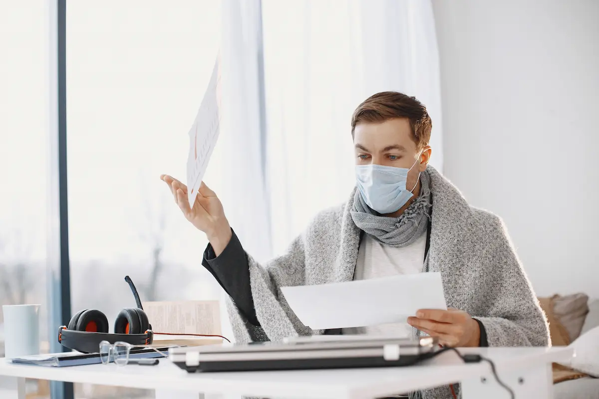Homme masqué en manteau et écharpe, tenant des feuilles à un bureau blanc, avec casque audio, lunettes et ordinateur portable