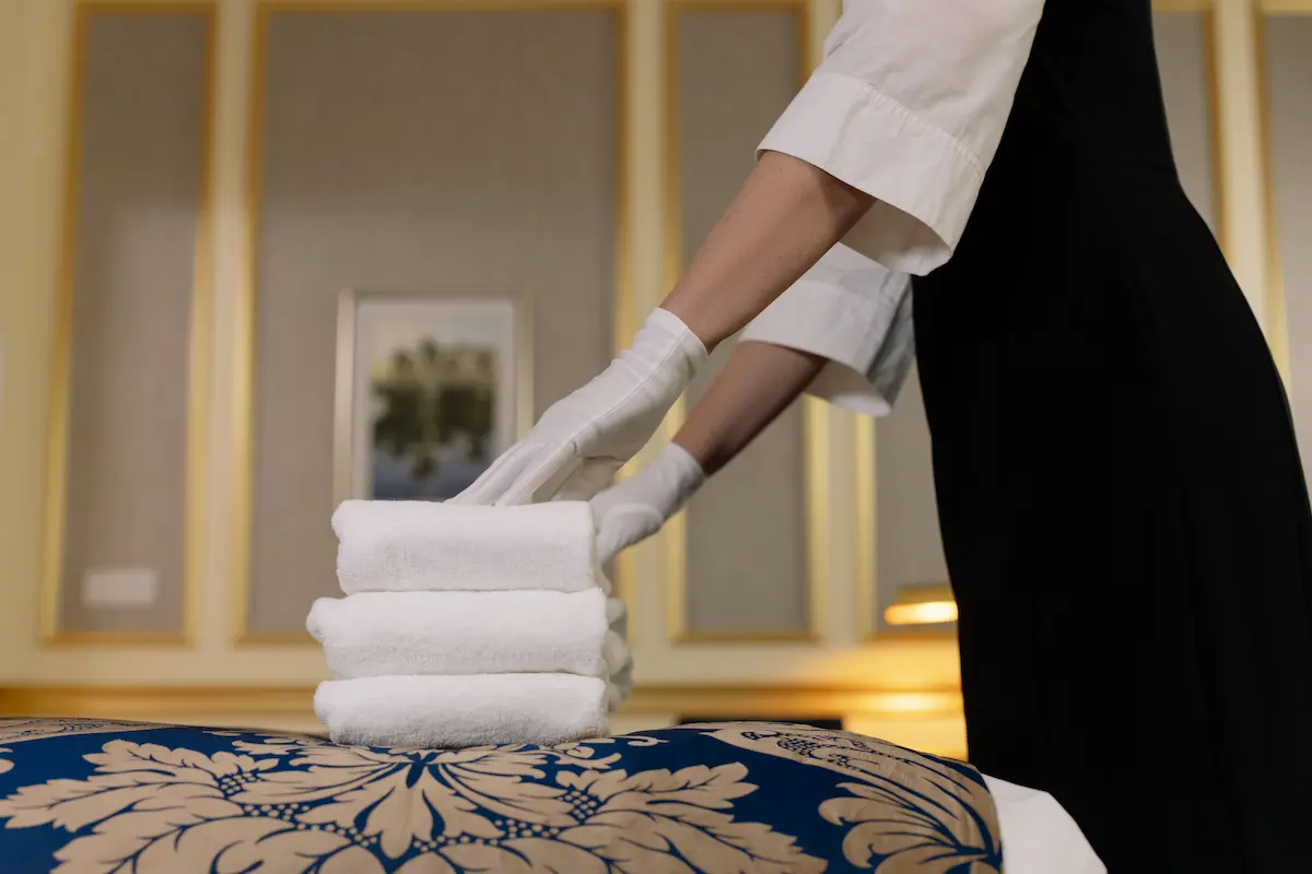 Employée d'étage en gants blancs déposant des serviettes pliées sur un lit d'hôtel